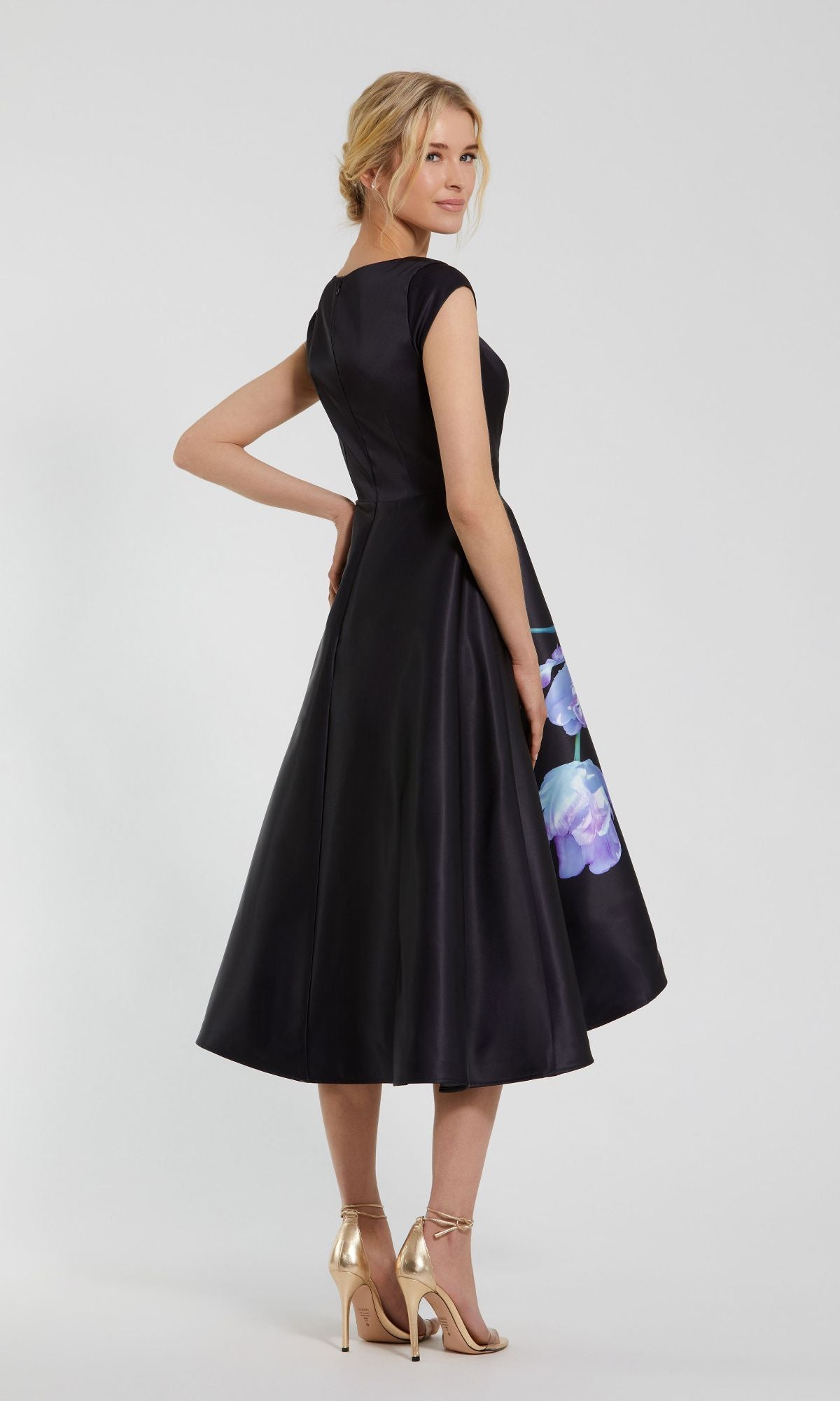A woman stands in profile with hand on hip and gold heels, wearing the Mac Duggal 20995 black satin wedding-guest dress featuring cap sleeves, a flared skirt, and floral accents.